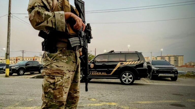Policial Federal armado, em, operação. Ao fundo uma viatura.
